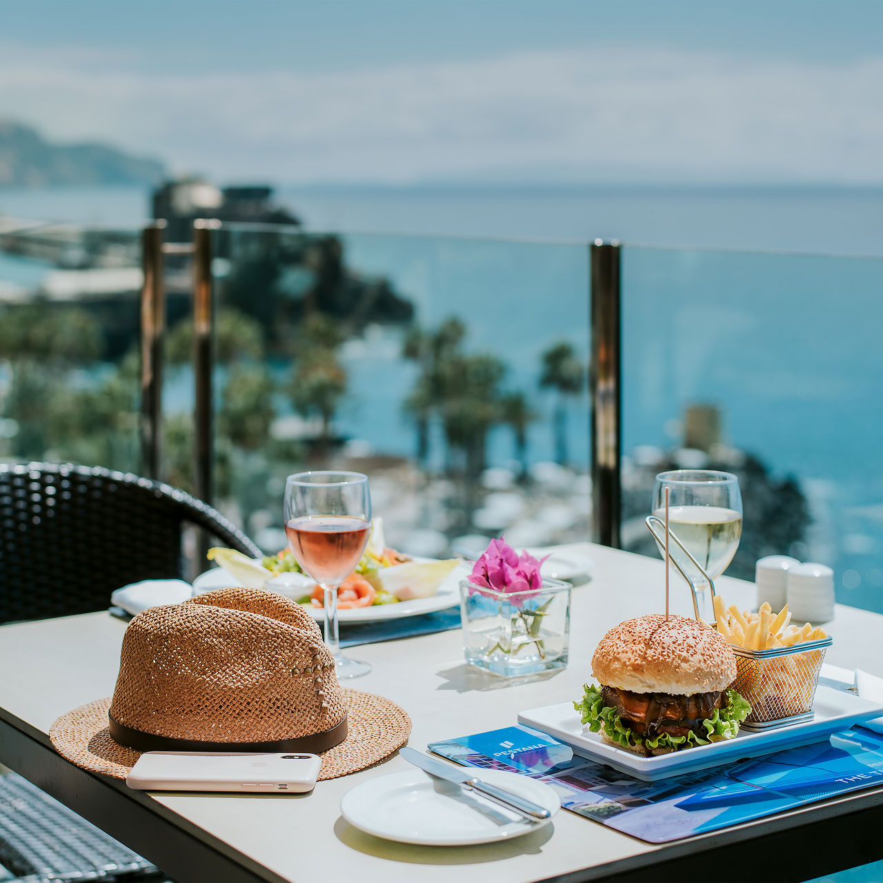 Table at Pestana Madeira Beach Club with a sea view, featuring hamburgers, fries, wine, and a hat