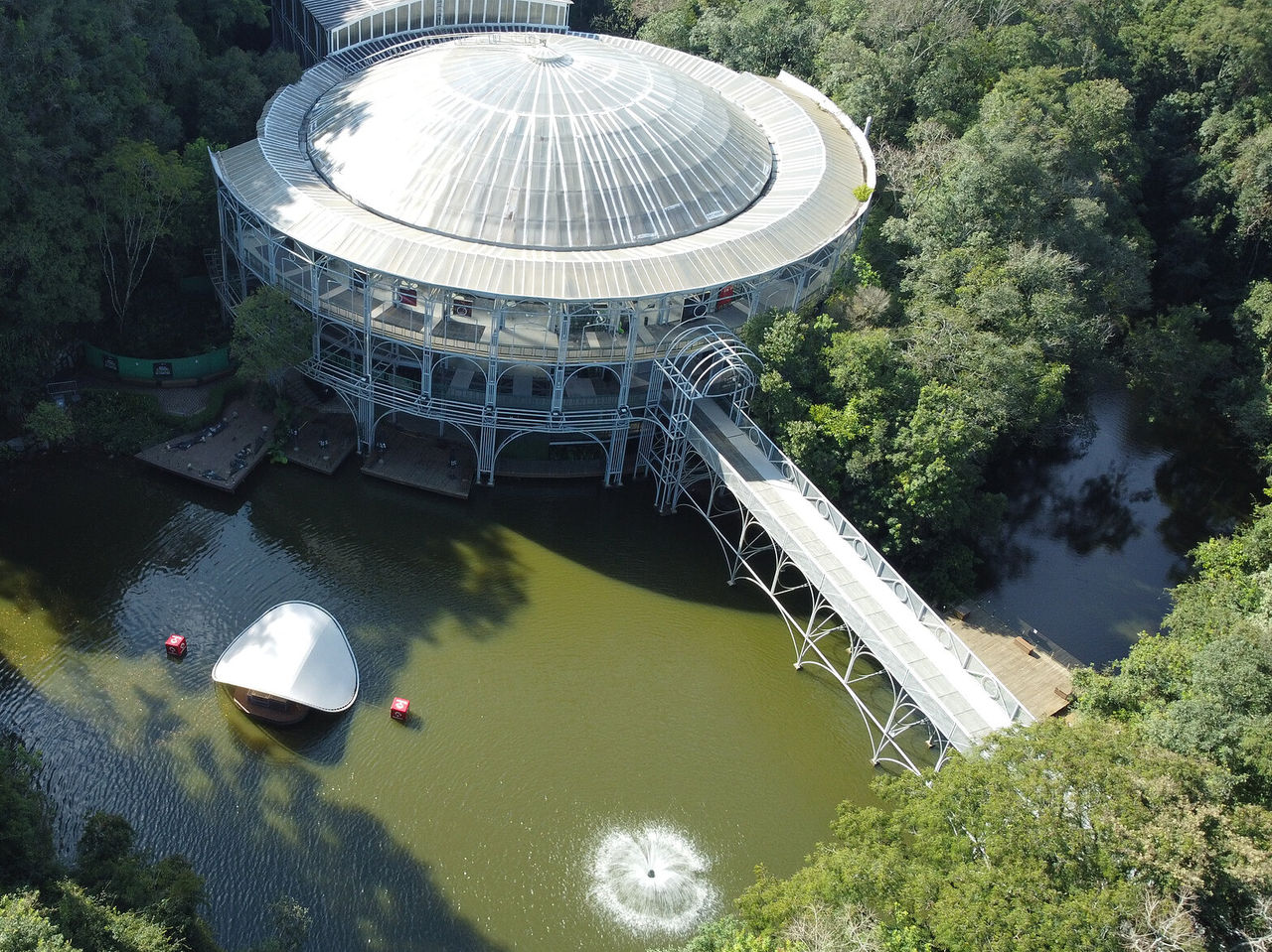 Aerial view of the Ópera de Arame, with its iconic round structure, surrounded by nature, with a bridge and a lake
