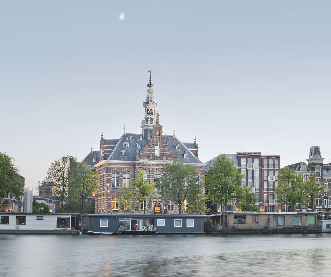 Amsterdam hotel overlooking a historic canal house beside calm water at dusk beneath a pale evening moon