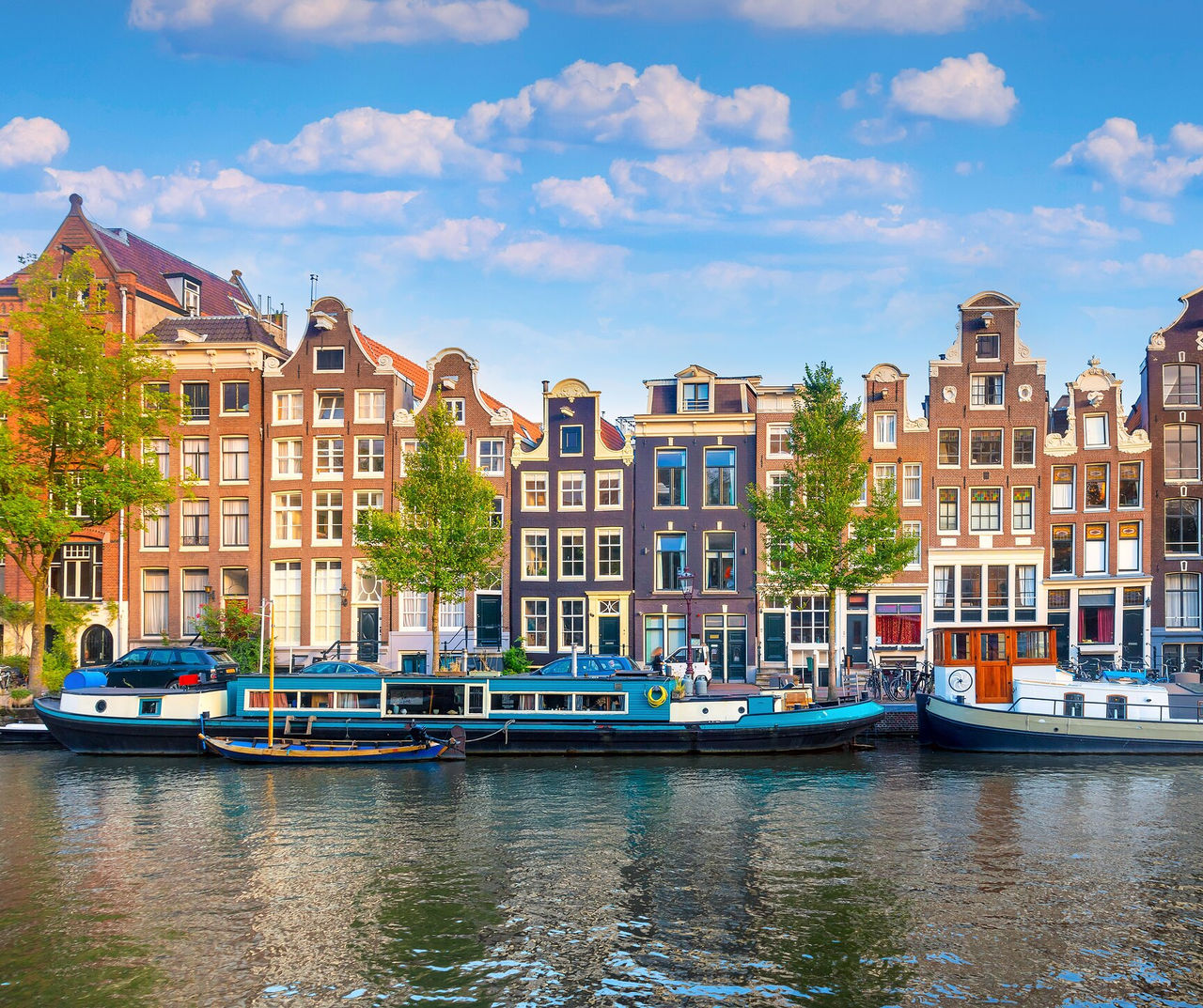 Amsterdam hotel featuring canal boats beside narrow brick townhouses beneath bright summer clouds and blue skies